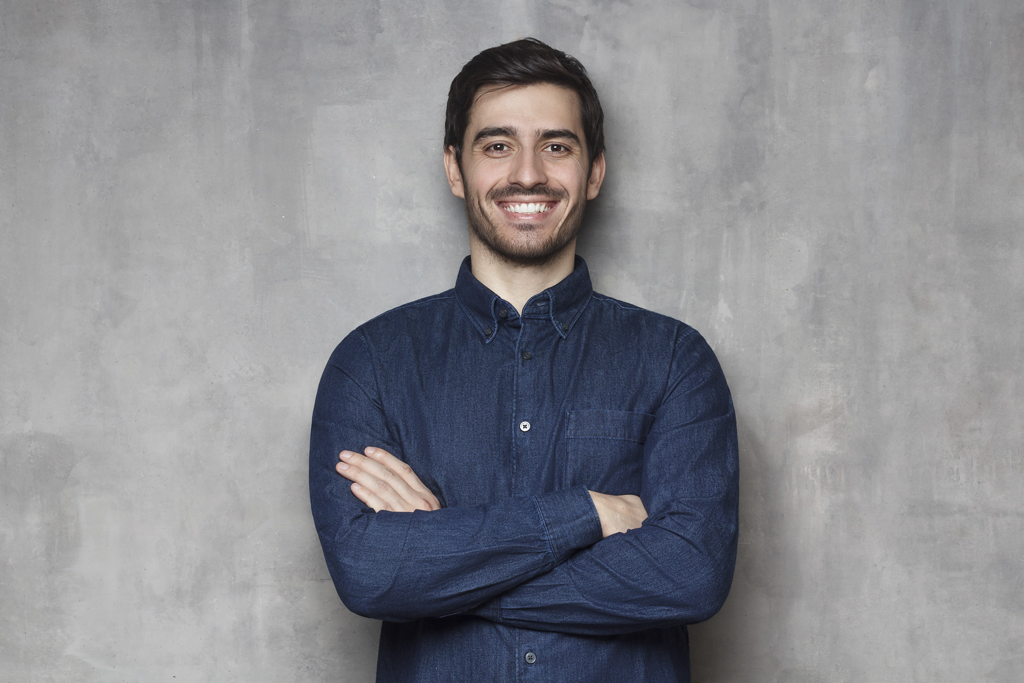 Trendy yound man standing with crossed arms and confident look against gray textured wall