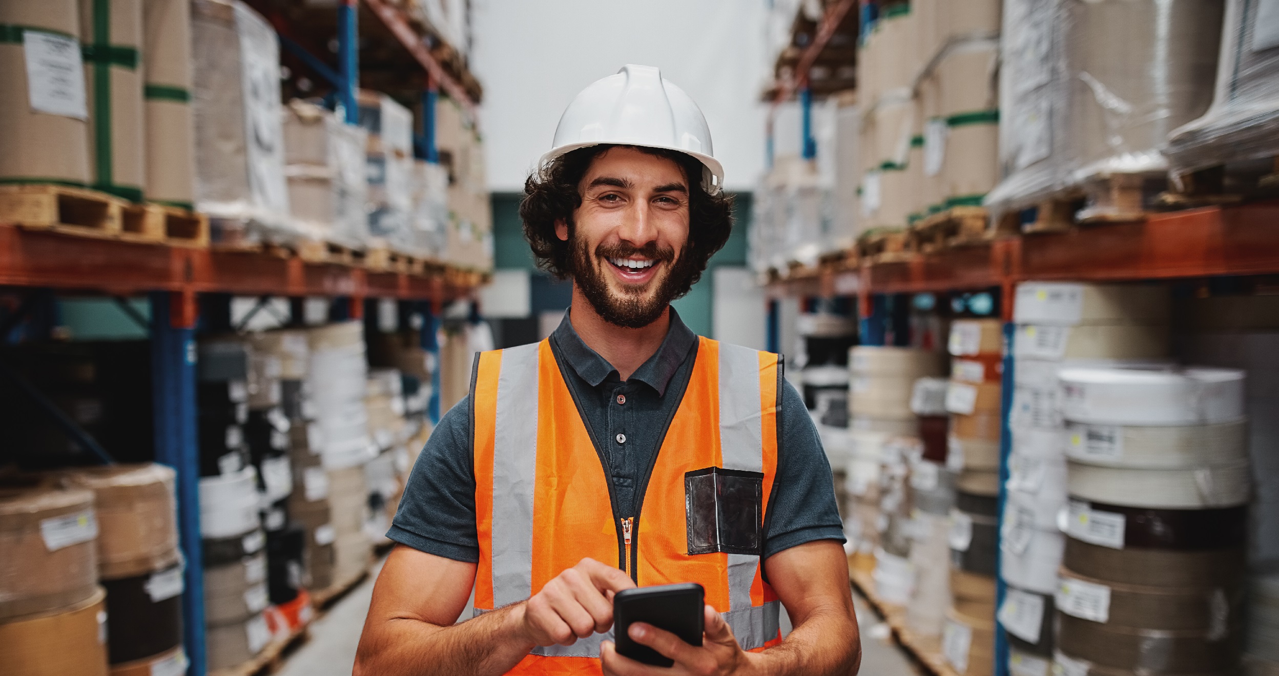 Young smiling male worker in warehouse wearing safety helmet and vest standing between shelf with goods using cellphone